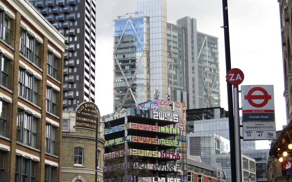 A city street scene in Shoreditch with tall modern high-rise buildings and older brick structures. Visible in the foreground is a parking sign, a bus stop sign, and a red and white bus directional sig